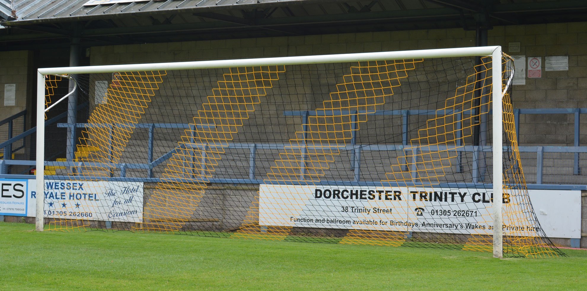 Ein Fußballtor mit gelb-schwarzem Netz vor leeren Tribünen im Dorchester Trinity Club Stadion.