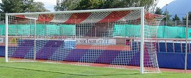Ein Fußballtor auf einem grünen Spielfeld mit bunter Tribüne im Hintergrund, umgeben von Bäumen und einem sonnigen Himmel.